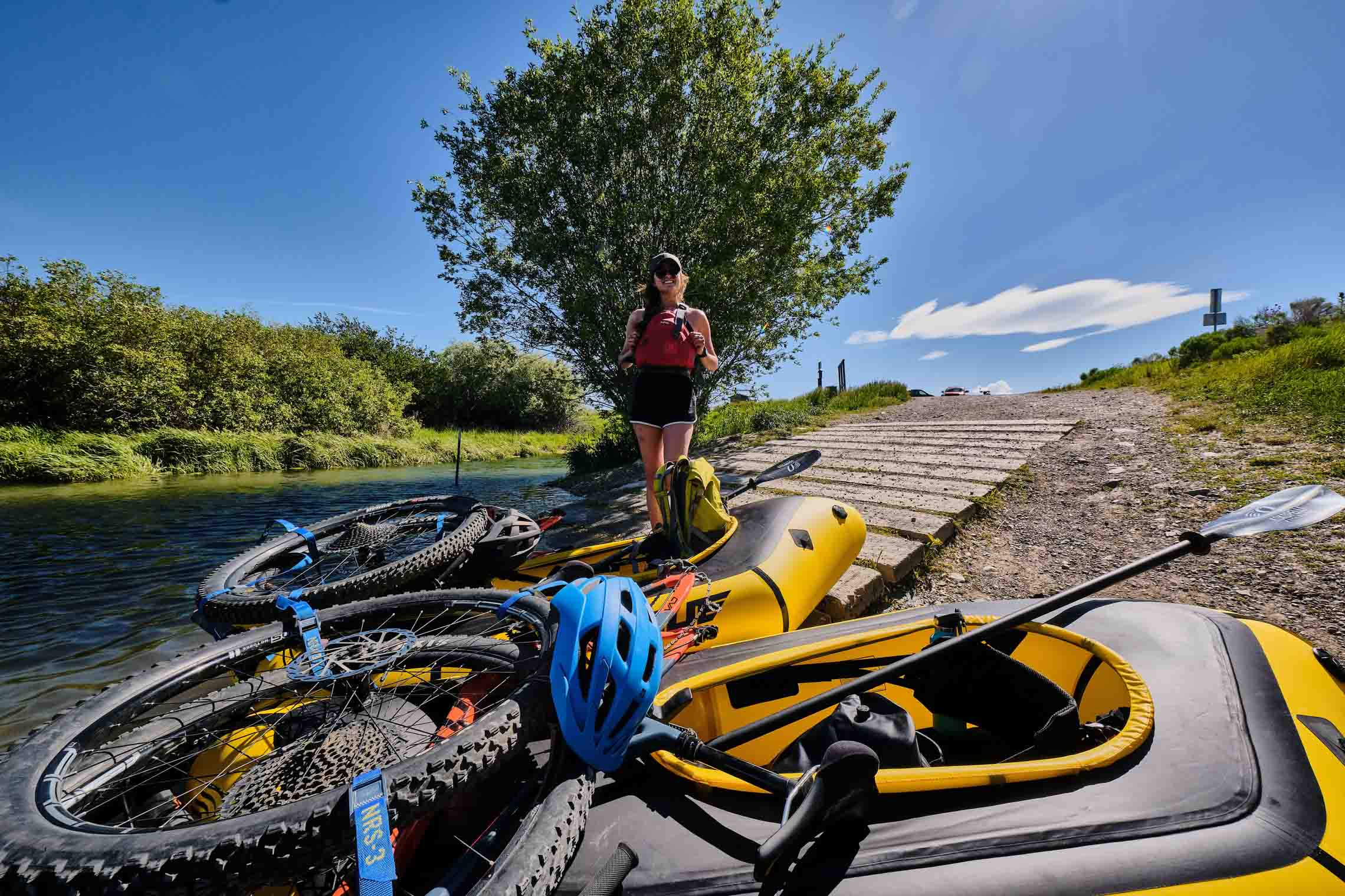 Bikes packed on packrafts next to water with a woman standing near them smiling