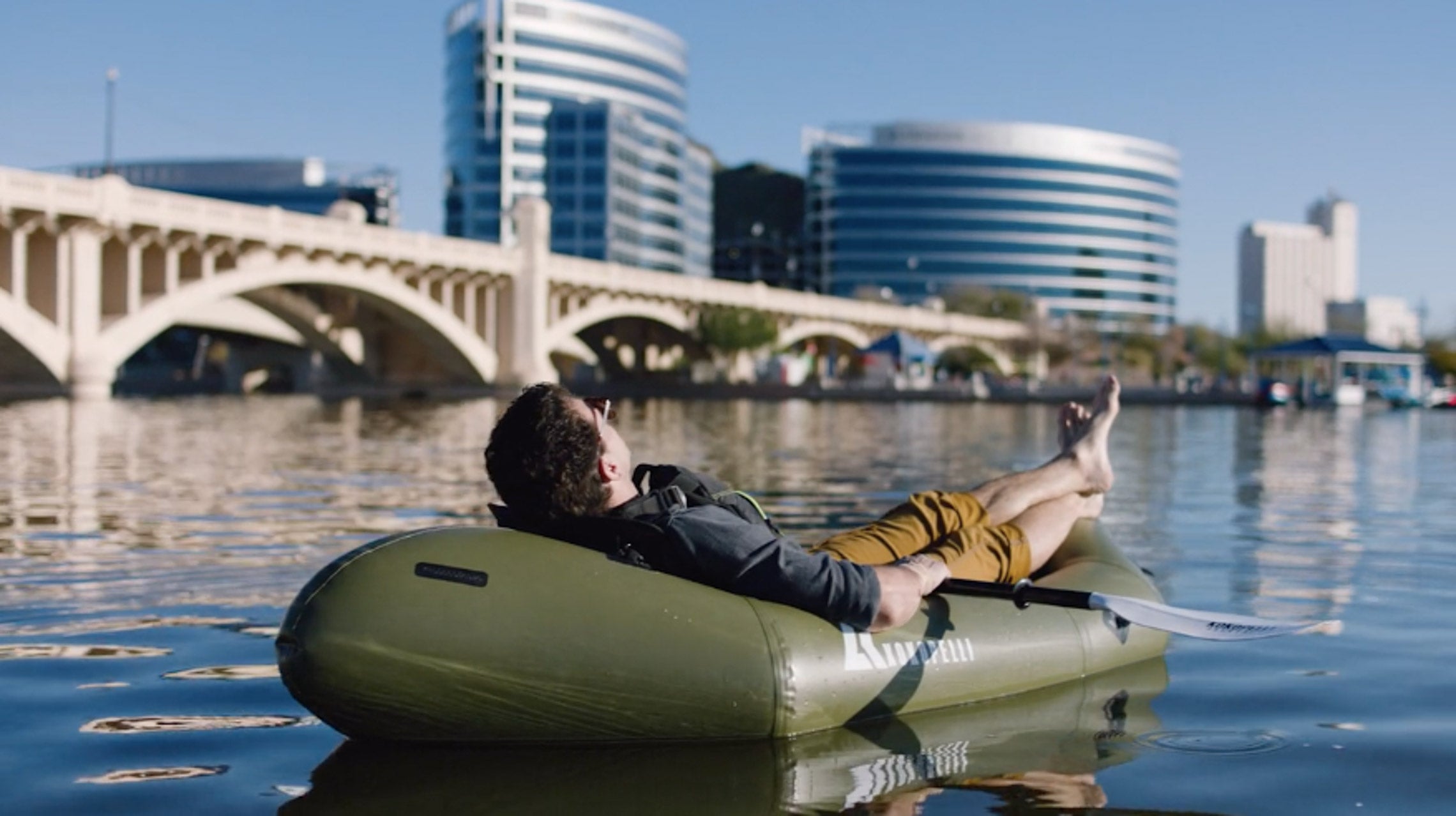 Man laying back in an XPD Kokopelli Packraft on water next to a city