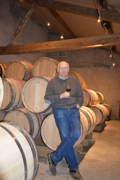 Winemaker François d'Allaines standing in front of wooden barrels in his cellar, holding a glass of red wine.