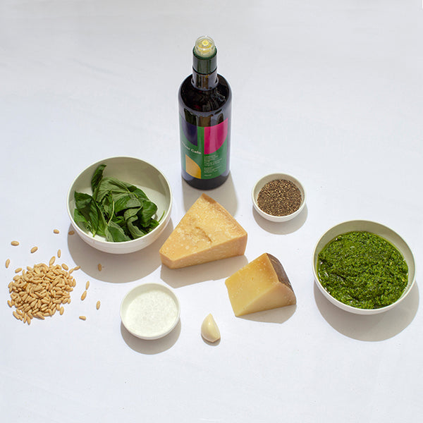 A bowl of basil leaves, a bowl of ground black pepper, a bowl of sea salt, a clove of garlic, some pine nuts, a wedge of parmesan, a wedge of pecorino, a bottle of River Cafe olive oil and a bowl of bright green pesto.