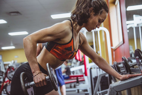 A brunette, muscular woman with a ponytail leans forward at a gym as she lifts an arm weight