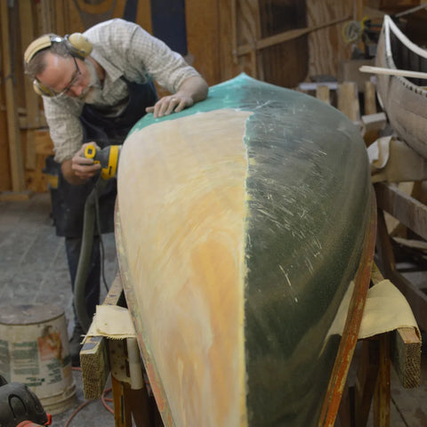 A man sands paint from a wooden canoe