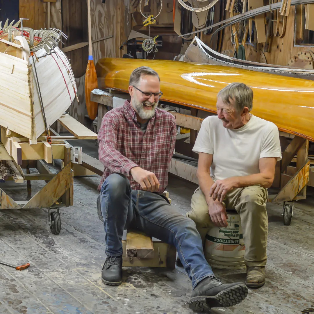 Two men sitting in a boat-building workshop, laughing together