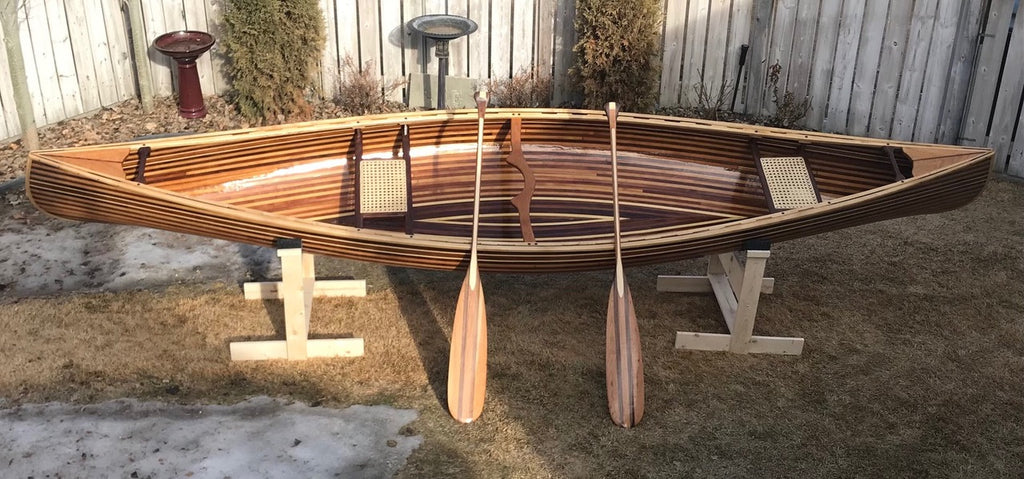 A wooden canoe with an alternating light-dark strip pattern sits outside in a cradle with two handmade paddles leaned against it