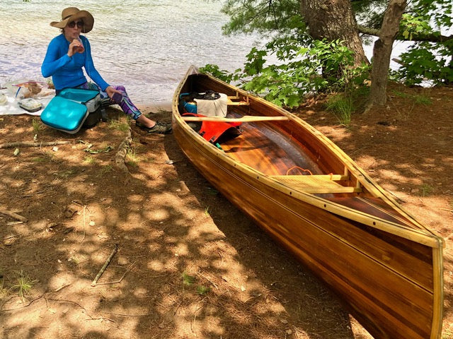 A woman sits by the shore sipping a drink, with a wooden canoe to her right