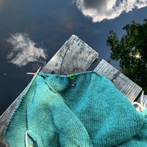 An aqua-colored knit in progress lies on a dock with a lake reflecting the sunny sky beneath it.