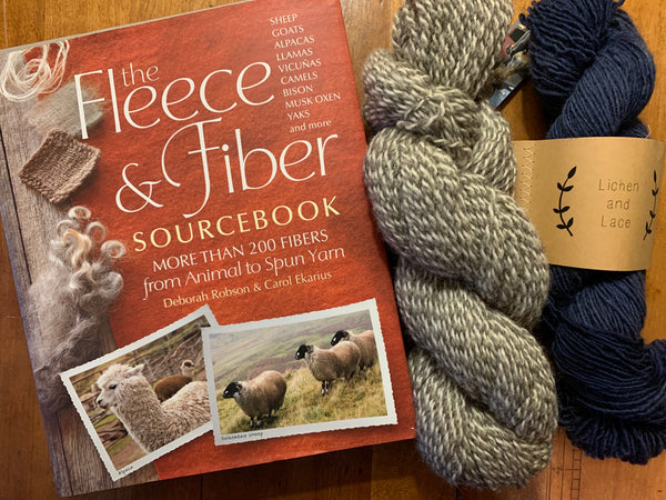 The Fleece & Fiber Sourcebook lies on a table next to a navy blue skein of yarn and a gray and white marled skein of yarn.