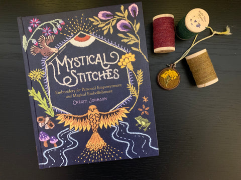 The book Mystical Stitches lies next to spools of wool thread in gold and cranberry, and a small wood embroidery hoop on a thread necklace, embroidered with a yellow flower design on gold wool fabric.