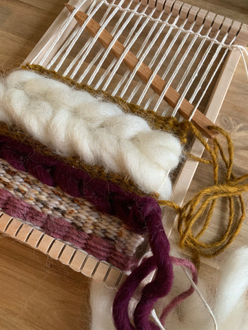 A wood tapestry needle is woven through a small hand loom along with yarns of mauve and gold, and a white braid of roving.