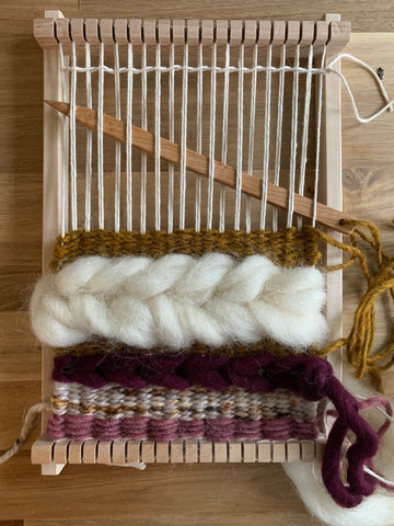 A small hand loom rests on a table with woven colors of mauve and gold, and a large white braid made of roving.