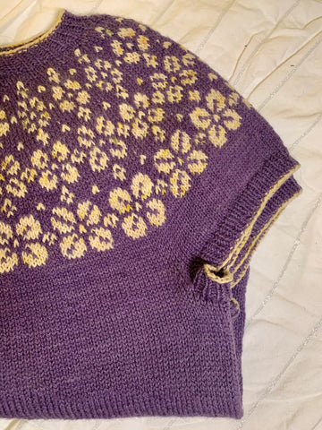A purple short sleeved wool sweater sits folded. It has a knit yoke of flowers in a white yarn speckled with yellow and green.