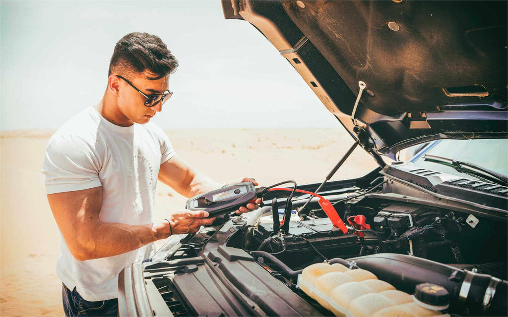 Man using portable jump starter on car battery in desert setting, illustrating smart motorist preparedness.