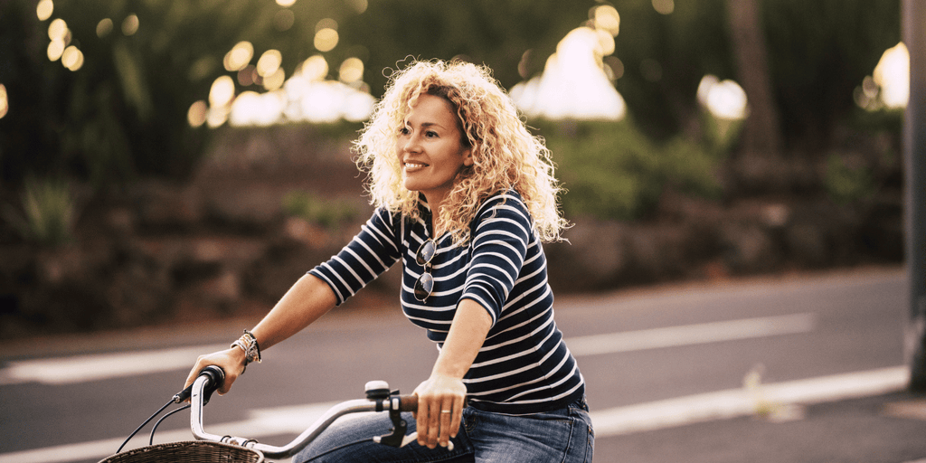 Beautiful woman with blond curly hair riding a bicycle and smiling!  wearing jeans and a t-shirt, out for a casual, fun ride on a sunny day with trees behind her.