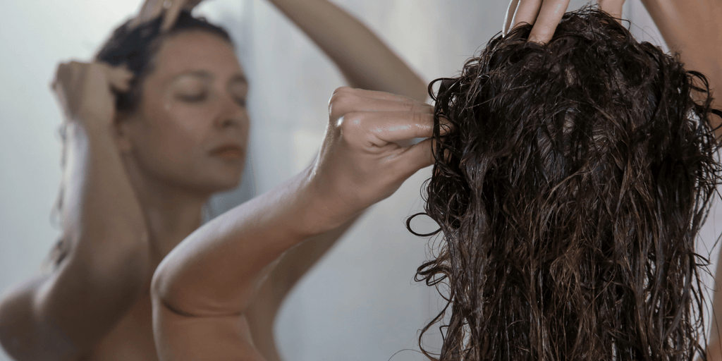 Woman with dark brown wet hair is massaging her scalp while looking in a mirror.  She is carefully massaging her scalp with her fingertips.