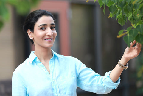 Apsara Skin Care founder Sheetal Rawal poses outside in a blue button down. She is smiling at the camera and reaching for some leaves on a branch.