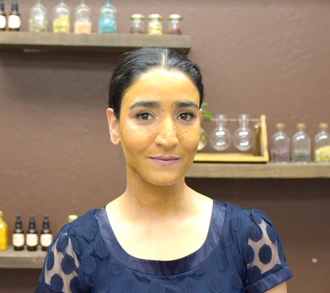 Apsara Skincare founder Sheetal Rawal wears a yellow face mask while standing in her office, with bottles of her skincare ingredients behind her.