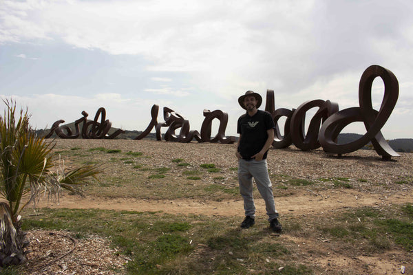 Touring Musician and Film Maker Kieran Wicks standing in Front of Large Metal Artwork Spelling Wide Brown Land, at Australian National Botanic Gardens Canberra. Living Art Lifestyle 