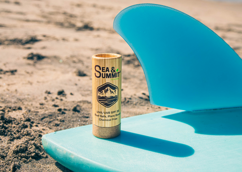 Sea & Summit's plastic-free sunscreen face stick leaning against a blue surfboard in the sand.