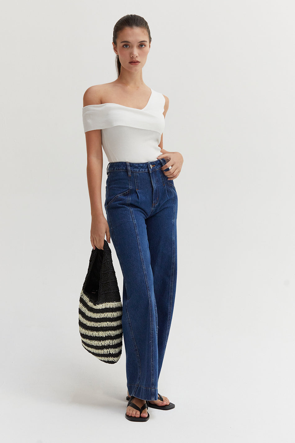 a model in dark luxe denim pants and a white off-shoulder-top with a striped bag