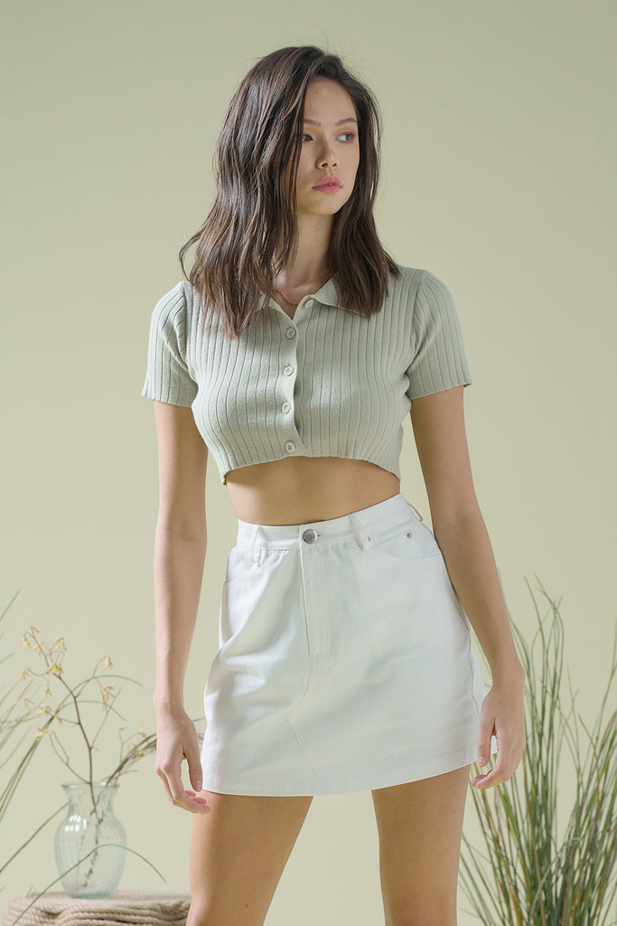 A woman wearing a sage cropped knit top and an ivory canvas mini skirt.