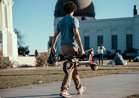 Person casually carrying a folded Kaabo electric scooter in an urban park, exemplifying ease of portability and urban lifestyle.