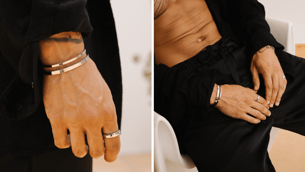 Man sat down with hands on his lap. He's wearing a timeless all black outfit and simple silver jewellery such as band rings and cuff bracelets