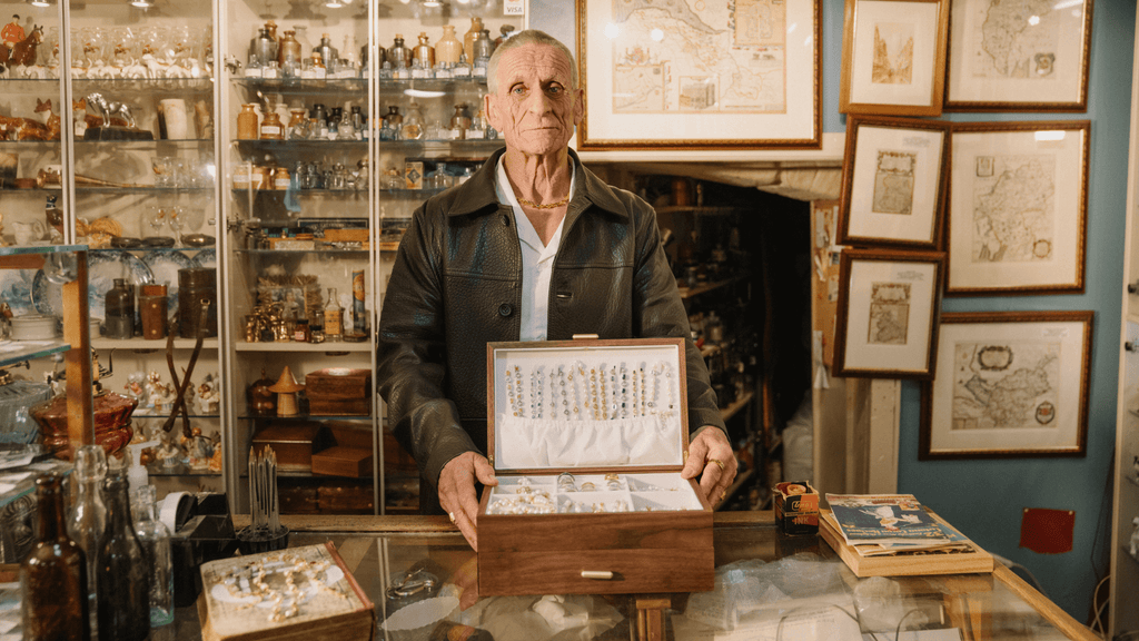 Man stood behind till of an antiques shop opening a men's jewelry box with loads of different men's jewelry including gold and silver rings, bracelets and necklaces