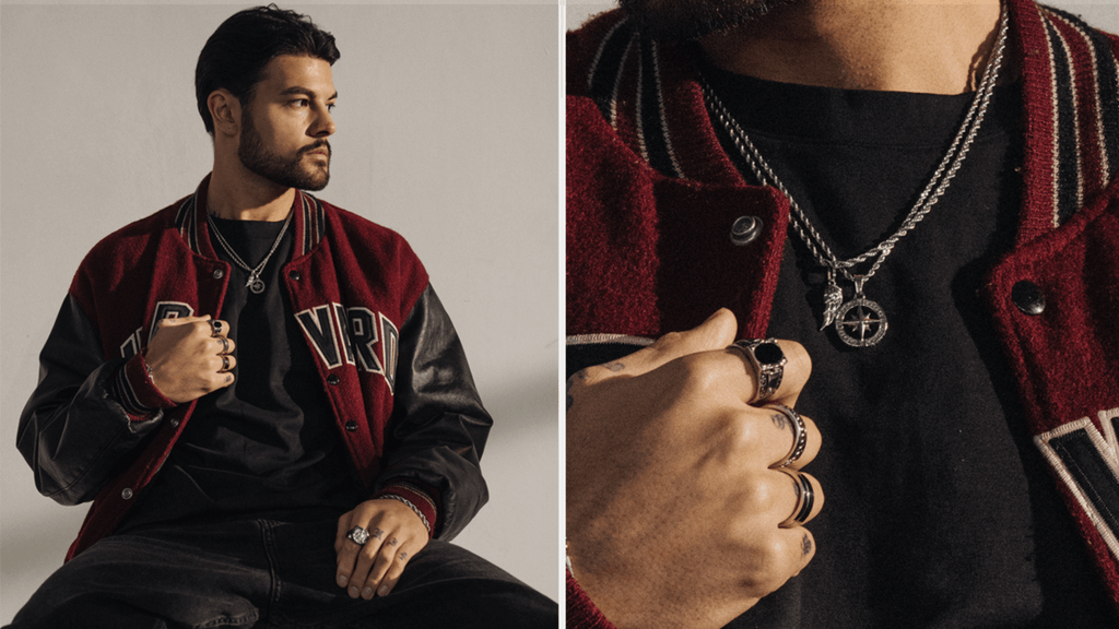 Man sat down wearing a casual red and black jacket. He's accessorising some silver jewelry including rings, bracelets and chains