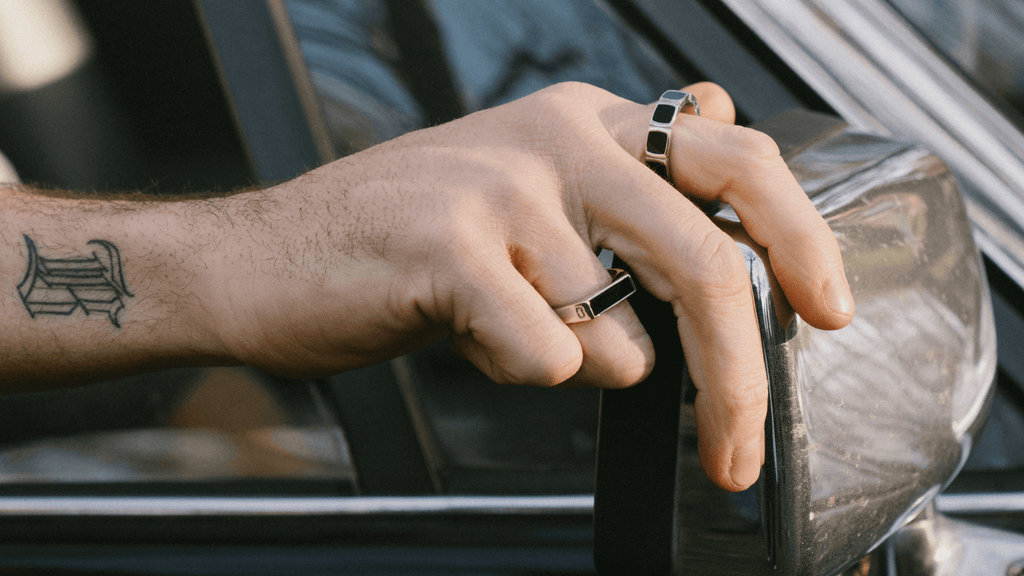 Man's hand adjusting a car side view mirror. He is wearing two sterling silver signet rings.