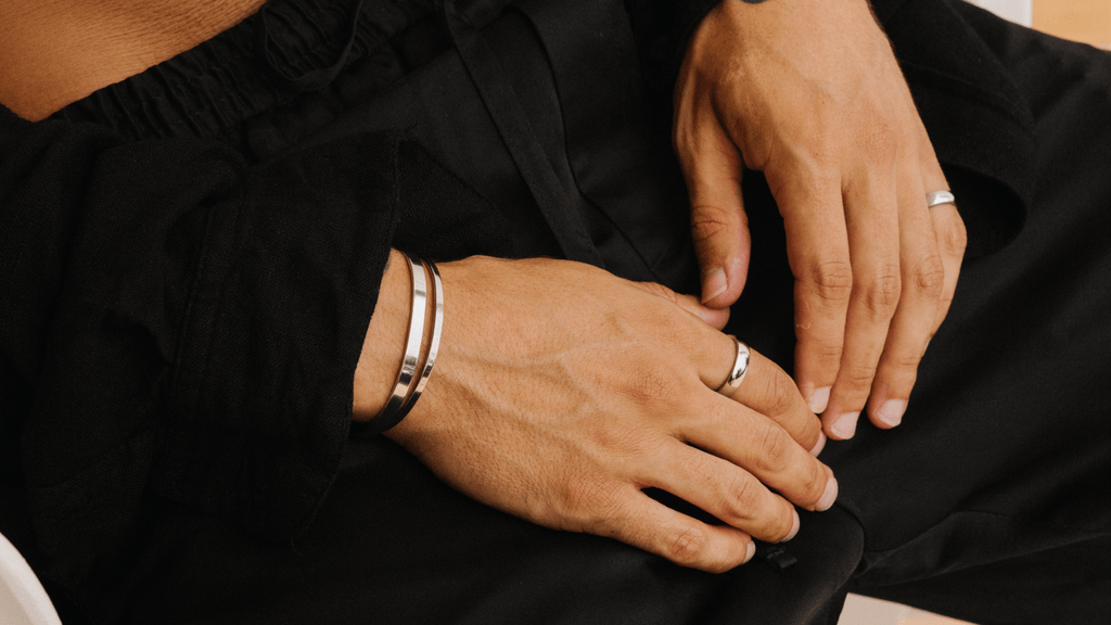Man's hands lay on his lap. He's wearing timeless black clothing and sleek cuff bracelets and rings in silver
