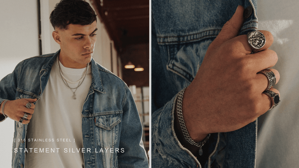 Man wearing a white t-shirt and blue denim jacket. He is stood leaning against a wall and modelling his stainless steel and silver jewellery including necklaces, rings an bracelets