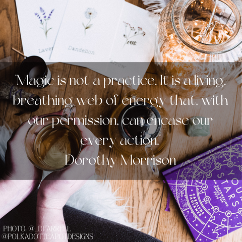 : A hand holding a small glass jar with a quote by Dorothy Morrison about magic being a web of energy. The background shows a wooden table with a book, a card, and a plant.