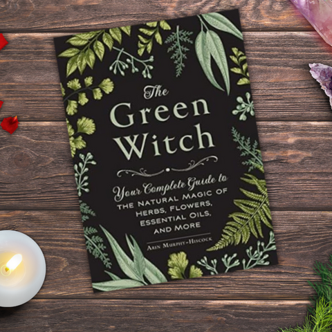 A photo realistic image of a book titled “The Green Witch” on a wooden table. The book is a guide to the natural magic of herbs, flowers, essential oils, and more by Arin Murphy-Hiscock.