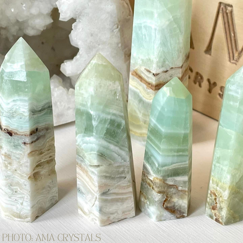 Four light green crystal towers with white and brown stripes on a white background. The crystals are in the shape of obelisks and are arranged from tallest to shortest. The image is from AMA Crystals, a company that sells natural and handmade crystal products.