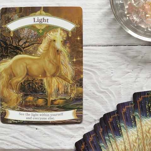 A photo of a tarot card titled “Light” that features a golden unicorn in a forest. The unicorn is standing in front of a tree with a golden aura and the card has a message about seeing the light within yourself and others.