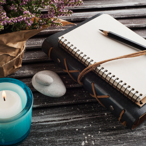 This is a photo of a notebook, a candle, and a bouquet of flowers on a wooden table. The notebook is black with a spiral binding and a brown elastic band around it.