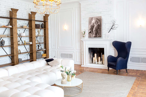photo of large white L-shaped sectional in the foreground and middle-ground with a white and gold coffee table in a white-walled room with ornamental plaster-work and white brick, wood floor, and gray area rug, and blue chair beside a white fireplace and large modern wooden bookshelf