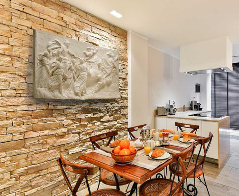 photo of a plaster cast sculpture relief of a woman flying and leading a chariot with a man pulled by horses towards the right while a cherub and other women fill the scene, on a tan brick wall with a kitchen in the background and a dining table with orange bowls in the foreground