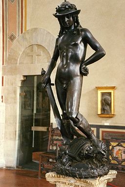 photo of bronze statue of male nude in hat and tall sandals, holding sword with other hand on hip, atop pedestal in off-white-colored gallery