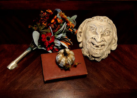 photo of plaster cast sculpture of grotesque faun face on a dark table next to fall flowers, a pumpkin, and a red book