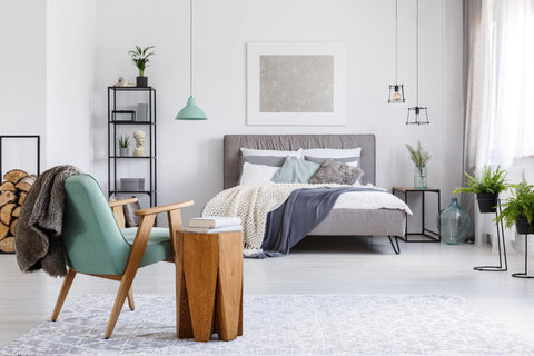 photo of bedroom with gray bed and headboard with white, blue, and mint-green blankets and pillows in background and mint-green and wooden modern chair and wooden side table in foreground and various plants, lamps, and other items