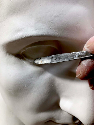 closeup of plaster cast of male bust showing an eye being worked with a sculpting tool