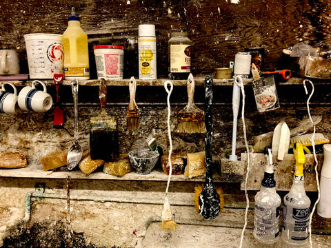 tool shelf with brushes, spoons, spray bottles, cans, jars, buckets and drinking mugs with plaster splattered on shelves and wall