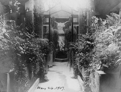 black and white photo of Victory of Samothrace sculpture at the end of a path bordered by plants in a glass-roofed conservatory
