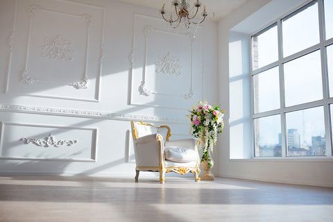 photo of ornate white and gold chair and tall urn with colorful flowers in the middle of a large room with wall window on the right and plaster-work on other visible wall and a crystal chandelier