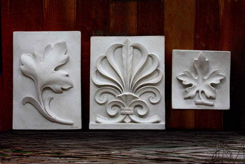 Set of Three ornamental plaster plaques, largest on the left and the smallest on the right, on a rustic background with warm toned wood and reeds below.