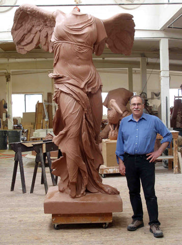 photo of Victory of Samothrace model in red clay beside sculptor Robert Shure in a white-walled and wooden-floored studio with various sculptures, saw horses, and tables