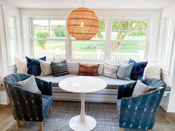 Kitchen nook with seating under a large window, decorated with custom grey cushion and a selection of vintage and new throw pillows. The kitchen nook is completed with a modern white round table and a blue accent chair on each end. 