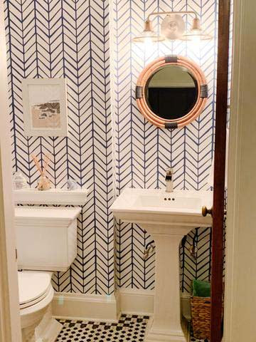 Small bright powder bathroom with patterned wall paper, round bamboo mirror and black and white floor tiles.