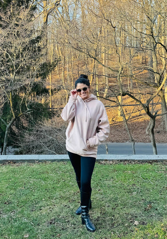 Woman wearing sunglasses with black boots, black leggings, and a pink hoodie with woods and grass behind her showing soccer mom outfit number 3